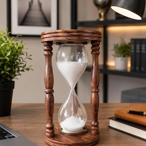 May include: An hourglass with a dark brown wooden frame featuring four turned pillars and circular top and bottom bases. The clear glass bulb contains white sand, with a portion currently flowing from the top chamber into the bottom chamber.