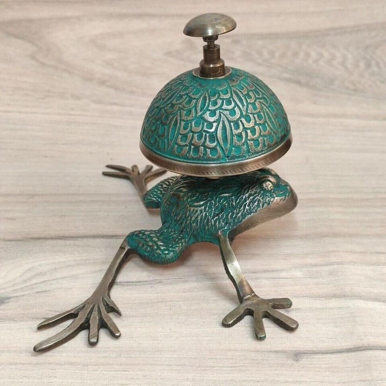May include: A metal desk bell shaped like a frog with a teal and bronze finish. The bell dome features an intricate embossed scale pattern, while the frog base has extended limbs and textured skin. A small round knob sits atop the dome.