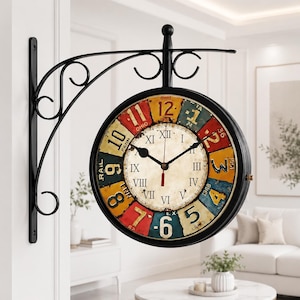 May include: A double-sided wall-mounted clock with a black metal scrollwork bracket. The clock face features a central cream dial with Roman numerals, surrounded by a colorful outer ring of segmented panels displaying Arabic numerals and text.