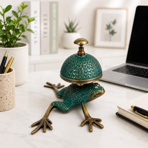 May include: A metal desk bell featuring a green frog base with a textured, scale-like pattern on its back and long, extended legs. The bell dome is a matching green with a decorative, repeating leaf-like pattern and a brass-colored top knob.