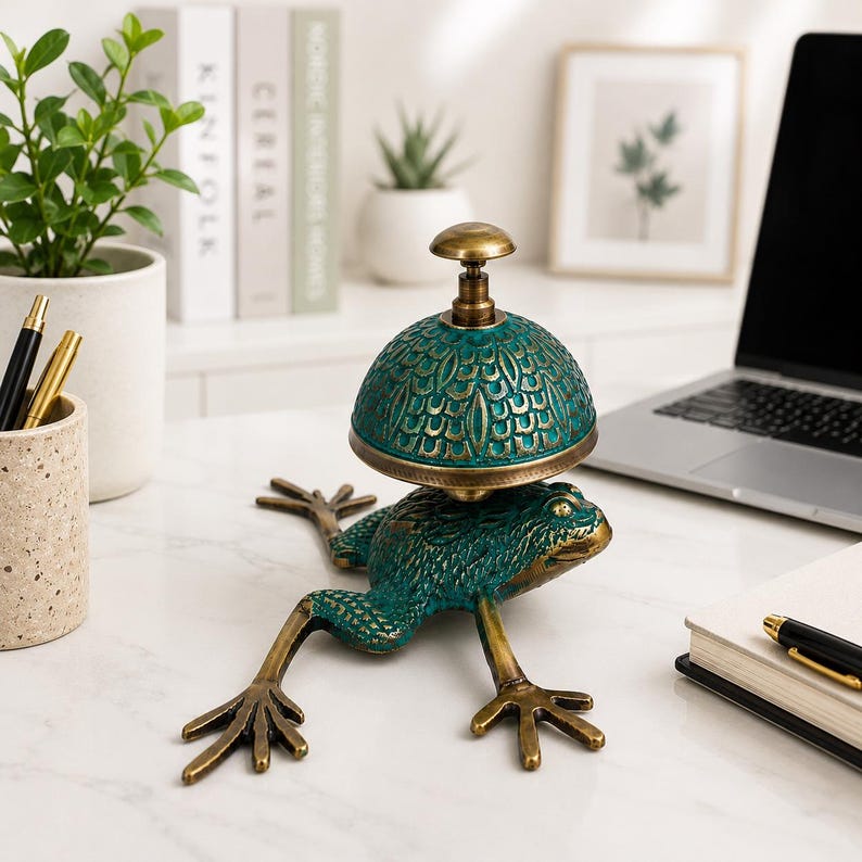 May include: A metal desk bell featuring a green frog base with a textured, scale-like pattern on its back and long, extended legs. The bell dome is a matching green with a decorative, repeating leaf-like pattern and a brass-colored top knob.