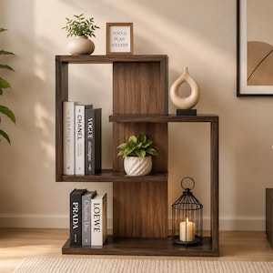 Könnte beinhalten: Ein dunkelbraunes Holzregal mit asymmetrischem, offenem Rahmendesign. Die Regale enthalten Bücher mit den Aufschriften Kinfolk, Chanel, Vogue, Prada und Loewe, eine kleine Topfpflanze, eine weiße Keramikvase, ein Schild mit Focus Plan Succeed und eine schwarze Laterne.