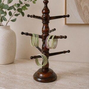 May include: A dark brown wooden jewelry stand with a central post and multiple horizontal arms. Several thin, glittery bangles in shades of light green and soft pink are hanging from the arms of the stand, which features a round base.