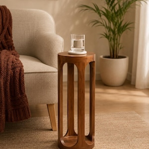Op de afbeelding: Een hoge, cilindervormige houten bijzettafel met een gladde, middenbruine afwerking. De tafel heeft een rond blad en een ronde voet die verbonden zijn door vier verticale, boogvormige steunen, wat zorgt voor een open en luchtige structuur. Een glas water staat bovenop.