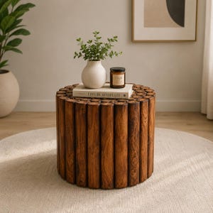 Puede incluir: Una mesa auxiliar cilíndrica construida con troncos de madera de color marrón oscuro dispuestos verticalmente en círculo. Un libro blanco con el texto 'LIVE BEAUTIFULLY' descansa encima, sosteniendo un jarrón de cerámica blanca y una vela.