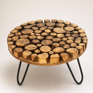 Pode incluir: Uma mesa redonda com um tampo construído a partir de numerosas pequenas fatias circulares de madeira de vários tamanhos e tons castanhos. A mesa é suportada por três pernas finas de metal preto em forma de gancho curvadas para fora.