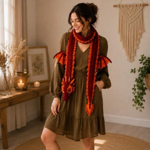 May include: A long, crocheted dragon scarf in shades of deep red and bright orange. The scarf features a detailed dragon head at one end, a pointed tail at the other, and two textured, wing-like appendages extending from the sides of the neck.