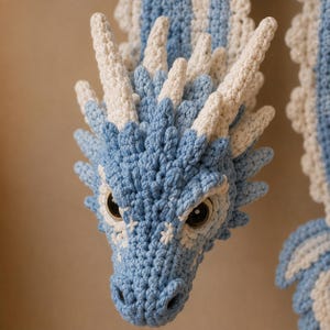 May include: A handcrafted crochet dragon head featuring light blue and cream yarn. The creature has prominent pointed horns, textured scales, and large dark inset eyes. The intricate stitching creates a detailed, layered appearance across the snout.