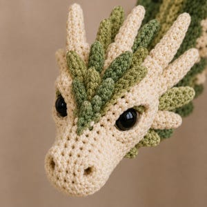 May include: A crochet dragon head featuring cream-colored yarn for the main structure and olive green yarn for the crest and accents. The creature has large, glossy black plastic eyes and a textured, stitched surface throughout its form.