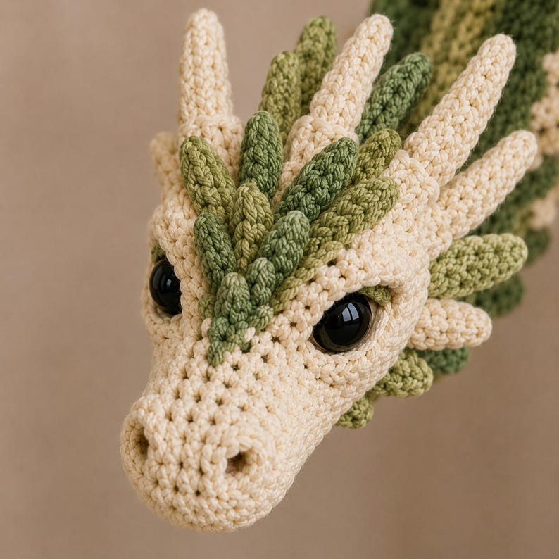 May include: A crochet dragon head featuring cream-colored yarn for the main structure and olive green yarn for the crest and accents. The creature has large, glossy black plastic eyes and a textured, stitched surface throughout its form.