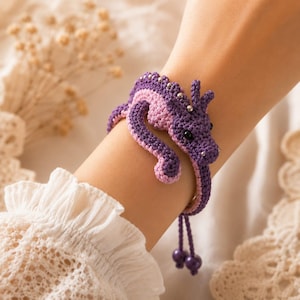 May include: A crochet bracelet featuring a purple and light pink dragon design. The dragon has a long, winding body, small silver beads along its back, and two dangling purple beads at the end of the adjustable cord closure.