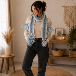 May include: A light blue and white crocheted dragon scarf featuring a detailed dragon head at one end, a pointed tail at the other, and two textured wings attached to the sides. The scarf has a scalloped white border along the edges.