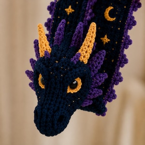 May include: A dark blue crocheted dragon scarf featuring purple side spikes and yellow horns. The scarf displays embroidered yellow stars and a crescent moon pattern along its length, with large, round, dark eyes set in yellow sockets.