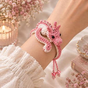 May include: A crochet bracelet featuring a pink and white dragon design with small silver beads along the spine, black bead eyes, and two dangling pink pearl-like beads at the ends of the adjustable cord closure.