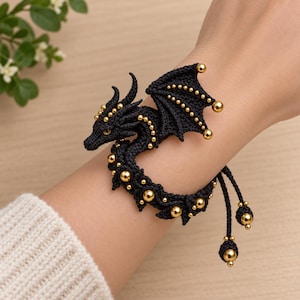Könnte beinhalten: Ein schwarzes Makramee-Armband in Drachenform mit goldfarbenen Perlen an Flügeln, Körper und Schwanz. Der Drache hat ein strukturiertes Knotendesign mit einem sichtbaren gelben Auge und zwei herabhängenden Schnüren mit goldenen Perlen.