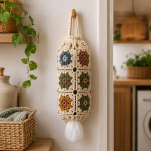 May include: A cylindrical crochet bag made of cream-colored yarn featuring a pattern of multi-colored granny squares in shades of blue, rust, olive green, and mustard. The bag has a top loop for hanging and an opening at the bottom for dispensing.
