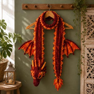 May include: A crochet dragon scarf in shades of deep red, orange, and yellow. The scarf features a detailed dragon head at one end, a pointed tail at the other, and two textured wings extending from the sides, all hanging from a wooden hanger.