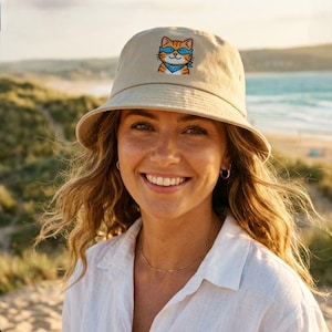 Peut inclure: Un chapeau bob beige orné d'une broderie représentant un chat tigré orange portant des lunettes de soleil bleues et un bandana bleu. Le chapeau présente un bord incliné vers le bas et une calotte structurée avec un motif cousu sur le devant.