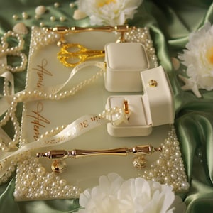 May include: A rectangular glass tray with gold handles, decorated with white pearls and the names Asmin and Adem with the date 30.05.2026. It features two cream-colored ring boxes, one containing a gold ring, and gold scissors on a ribbon.