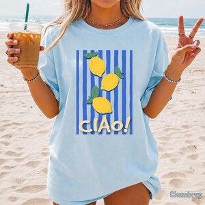 May include: A light blue t-shirt featuring a graphic print of three yellow lemons with green leaves arranged vertically over a pattern of dark blue vertical stripes. The text "CIAO!" is printed in a light yellow font below the lemons.