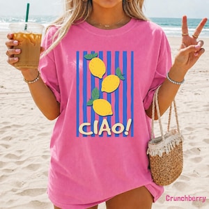 May include: A bright pink short-sleeve t-shirt featuring a graphic print of three yellow lemons with green leaves centered over vertical blue stripes. The text "CIAO!" is printed in a bold, light-colored font below the lemon illustration.