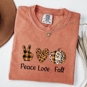 May include: A terracotta-colored short-sleeve t-shirt featuring a graphic print with a black and orange plaid peace sign, a leopard print heart, and a pumpkin with plaid and leopard patterns. The text reads Peace Love Fall in black.