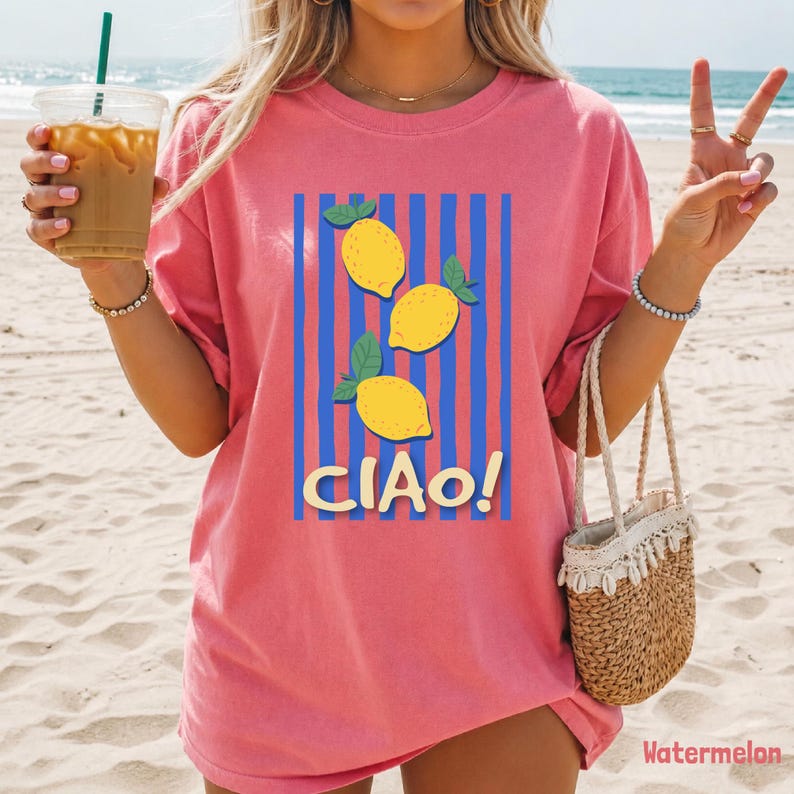 May include: A coral pink short-sleeve t-shirt featuring a graphic print of three yellow lemons with green leaves centered over vertical blue stripes. The text "CIAO!" is printed in a bold, cream-colored font below the lemon illustration.