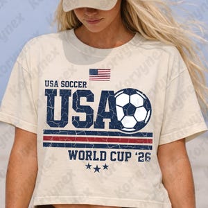 May include: A cream-colored short-sleeve t-shirt featuring a distressed graphic with the text USA SOCCER, USA, and WORLD CUP '26. The design includes a soccer ball, a small American flag, three stars, and horizontal red and blue stripes.