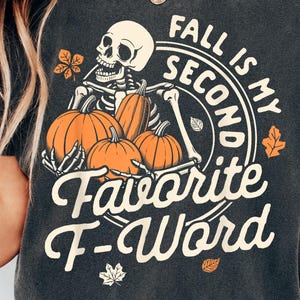 May include: A charcoal gray short-sleeve t-shirt featuring a graphic of a skeleton holding three orange pumpkins. The text reads "FALL IS MY SECOND FAVORITE F-WORD" in a cream-colored font, accented with small orange and white autumn leaf illustrations.