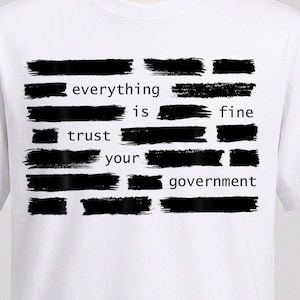 Op de afbeelding: Een wit t-shirt met korte mouwen en een afbeelding van zwarte horizontale penseelstreken. De tekst "everything is fine trust your government" is in zwarte kleine letters in het midden gedrukt en wordt gedeeltelijk bedekt door de penseelstreken.