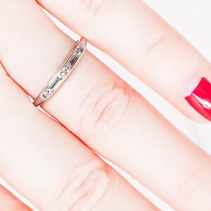 May include: A thin silver band with small diamonds set in a row. The ring is worn on a finger with red nail polish.