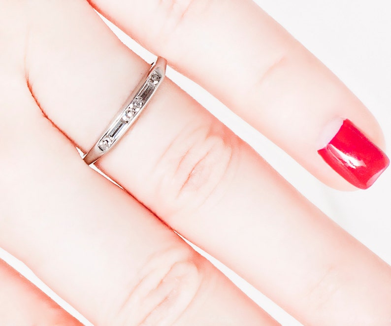 May include: A thin silver band with small diamonds set in a row. The ring is worn on a finger with red nail polish.
