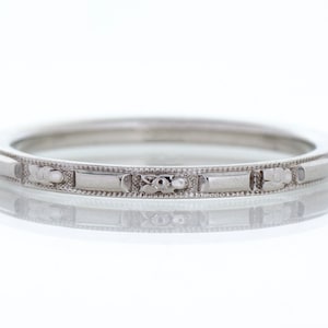 May include: A silver ring with a repeating pattern of rectangular and floral designs. The ring is thin and has a polished finish, reflecting light. The ring is likely made of a precious metal, such as silver or platinum.
