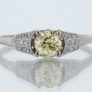 May include: A platinum engagement ring featuring a large, round, light yellow diamond center stone. The band is adorned with smaller diamonds and decorative filigree detailing on the shoulders. The ring is set against a reflective white background.