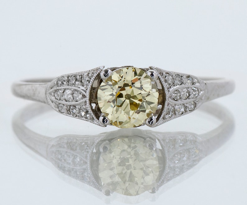 May include: A platinum engagement ring featuring a large, round, light yellow diamond center stone. The band is adorned with smaller diamonds and decorative filigree detailing on the shoulders. The ring is set against a reflective white background.