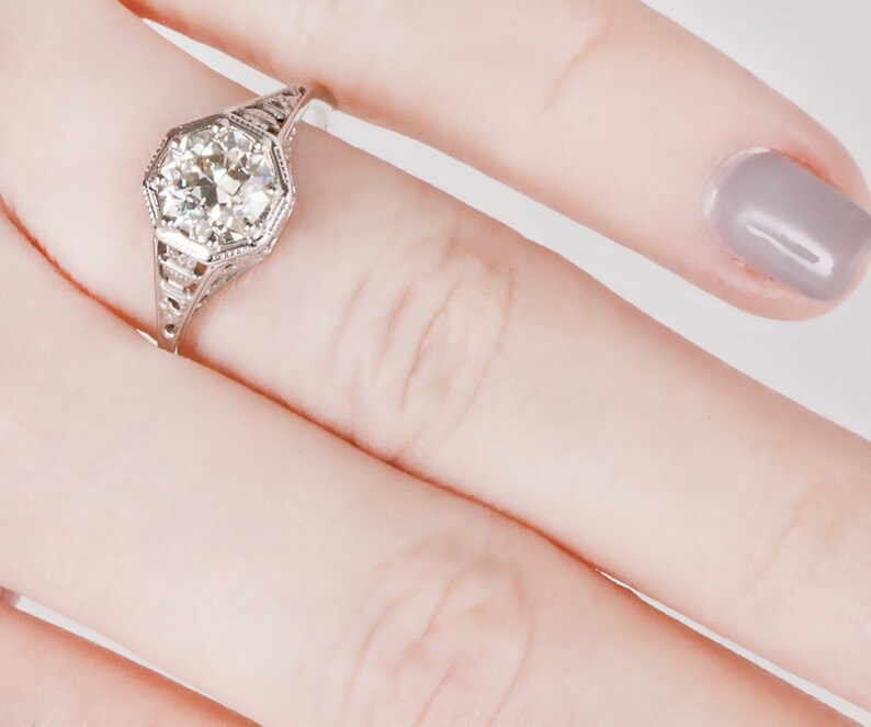 K&ouml;nnte beinhalten: Ein silberner Ring mit einem gro&szlig;en, runden, klaren Diamanten, der in einem sechseckigen Rahmen gefasst ist. Der Ring befindet sich an einem Finger mit grauem Nagellack.