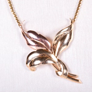 May include: A gold-toned necklace featuring a stylized leaf design. The pendant is composed of multiple overlapping leaves with intricate detailing, suspended from a delicate chain. The necklace has a polished finish, reflecting light.