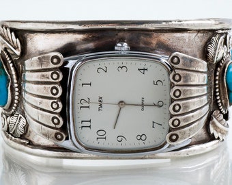 Puede incluir: Un reloj de plata con una esfera cuadrada y piedras turquesa a cada lado. El reloj está engastado en un brazalete de plata con un diseño nativo americano.