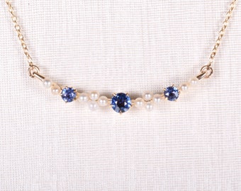 May include: A delicate gold necklace featuring a curved row of small, white pearls. Three vibrant blue gemstones are evenly spaced among the pearls. The necklace is displayed against a white, textured background.