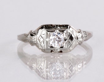 May include: A vintage engagement ring with a single round diamond set in a platinum band. The band is decorated with intricate filigree details.
