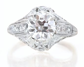 May include: A platinum ring with a large, round-cut diamond as the centerpiece. The ring features intricate filigree and smaller diamonds set along the band, creating a vintage or antique aesthetic. The ring is set against a white background.