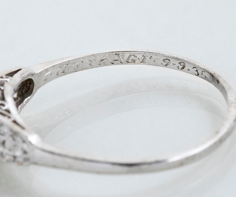 May include: Close-up of a silver ring with a filigree design and small gemstones. The ring is engraved with the text "AGT: 9-9-35". The ring is set against a white background.