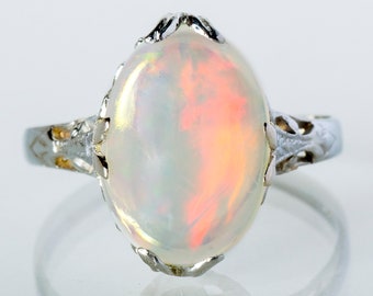 May include: An oval opal gemstone ring with a silver band. The opal has a milky white base with flashes of orange and yellow. The silver band features intricate filigree detailing on the shoulders of the ring.