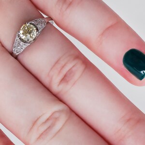 May include: A platinum ring featuring a central, round, light yellow diamond, flanked by smaller, clear diamonds. The ring is set on a finger with a dark green nail polish.