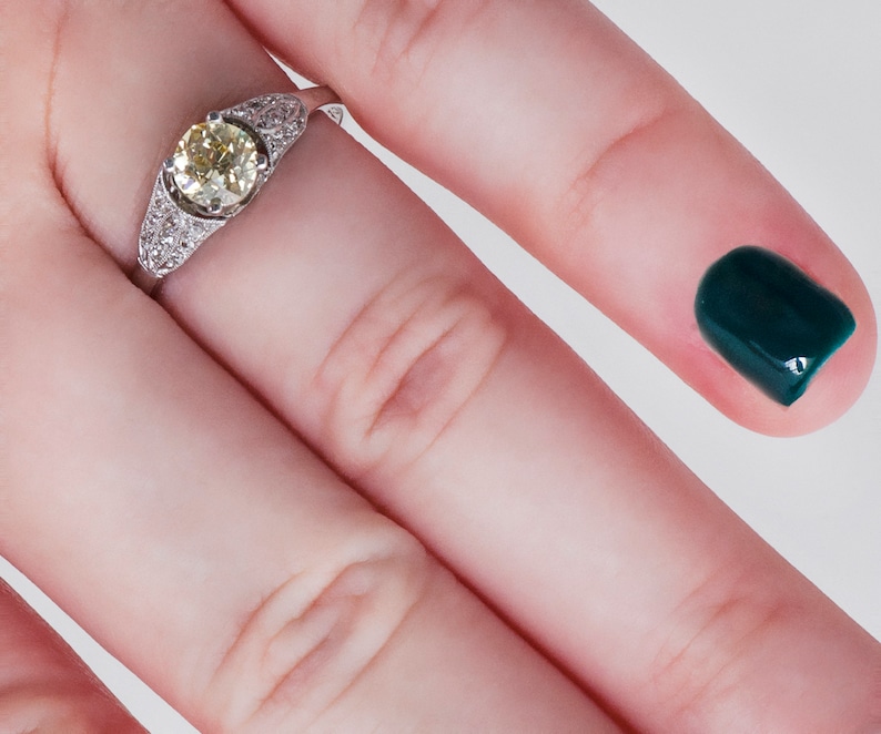 May include: A platinum ring featuring a central, round, light yellow diamond, flanked by smaller, clear diamonds. The ring is set on a finger with a dark green nail polish.