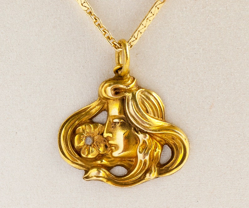 May include: A gold pendant necklace featuring a detailed Art Nouveau style design of a woman's face with flowing hair and a flower.