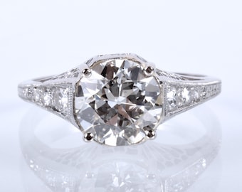 May include: A platinum engagement ring with a large, round-cut diamond as the centerpiece. Smaller diamonds are set along the band, with intricate filigree detailing on the shoulders. The ring is displayed on a reflective surface.