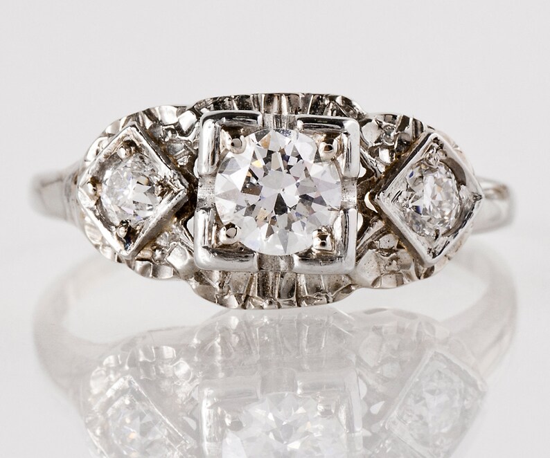 Puede incluir: Un anillo de plata vintage con un gran diamante redondo en el centro y dos diamantes cuadrados m&aacute;s peque&ntilde;os a cada lado.