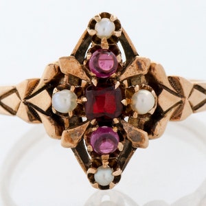 Puede incluir: Anillo dorado con una piedra roja rectangular central, rodeada de piedras rojas y blancas redondas m&aacute;s peque&ntilde;as. El anillo presenta un dise&ntilde;o geom&eacute;trico ornamentado en la banda y alrededor de las piedras centrales, creando una est&eacute;tica vintage.