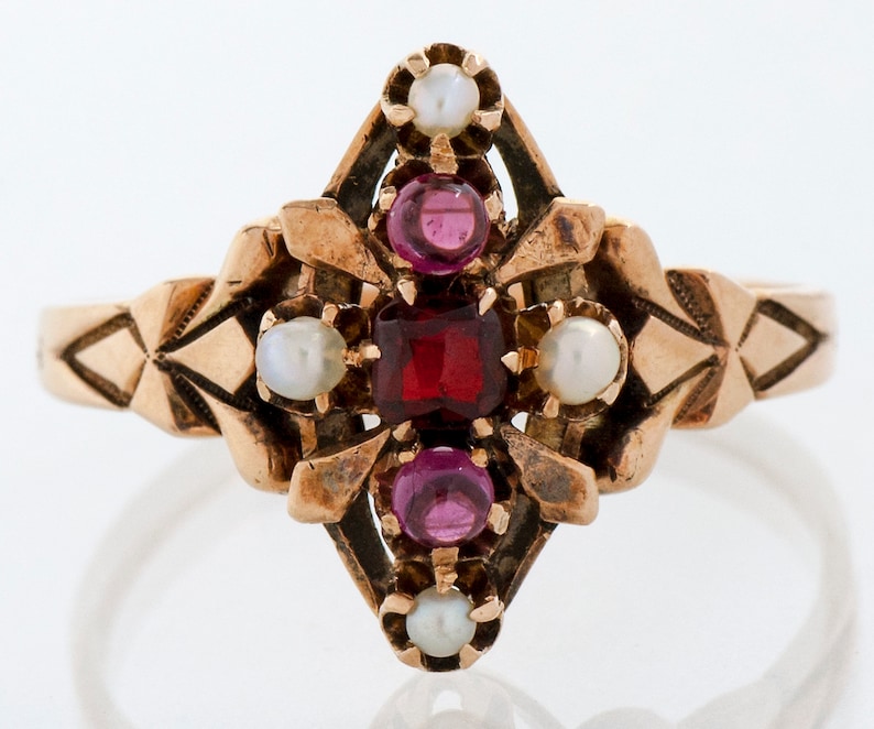 Puede incluir: Anillo dorado con una piedra roja rectangular central, rodeada de piedras rojas y blancas redondas m&aacute;s peque&ntilde;as. El anillo presenta un dise&ntilde;o geom&eacute;trico ornamentado en la banda y alrededor de las piedras centrales, creando una est&eacute;tica vintage.